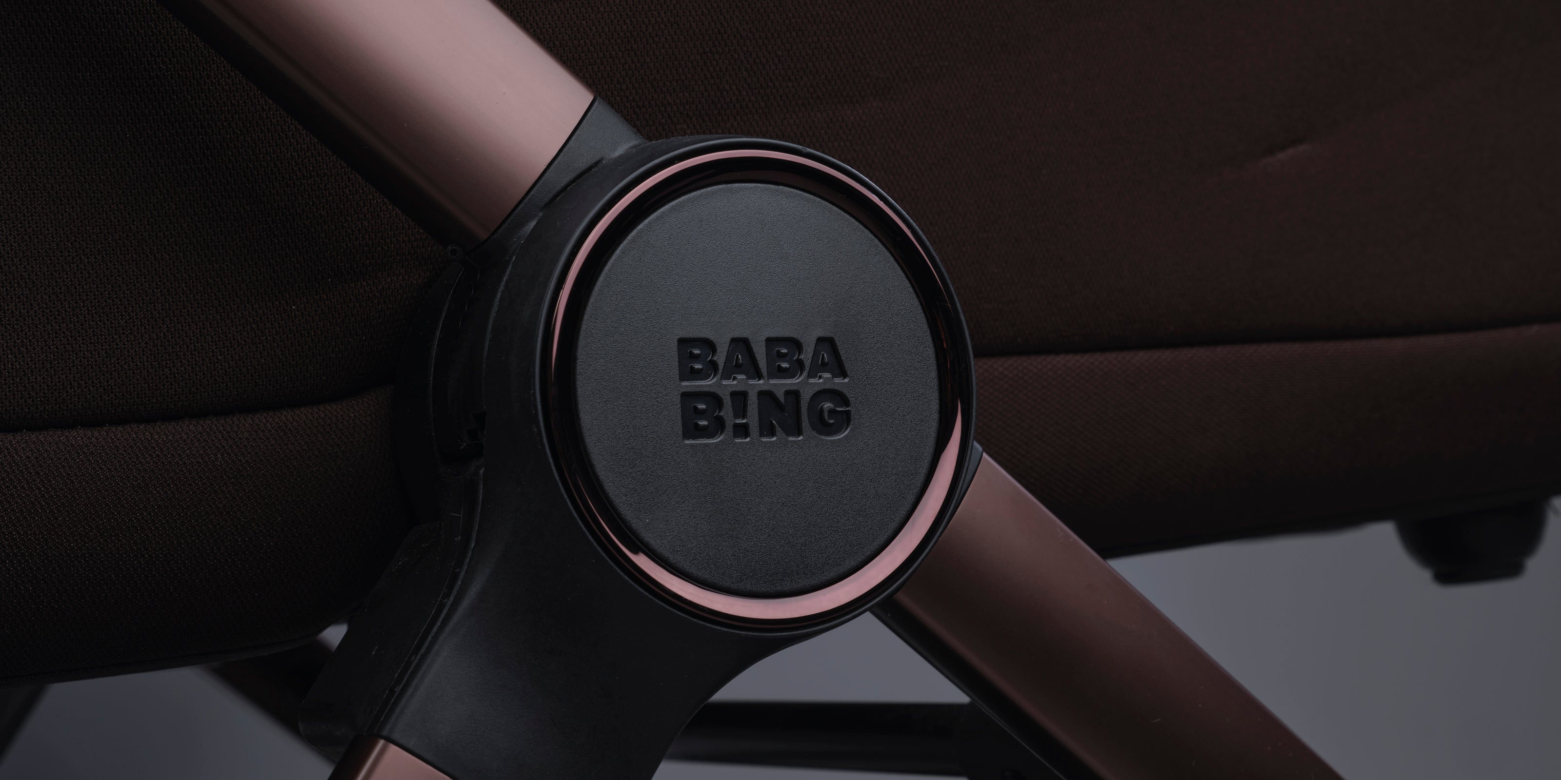 Close-up of a stroller frame with a black central hub featuring the embossed brand name 'BABA BING' and a metallic rose gold finish, set against dark brown stroller fabric.