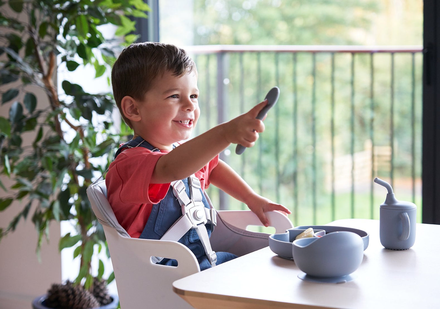 Smiling toddler in a high chair enjoying a meal at the table with natural light and toddler-safe utensils.