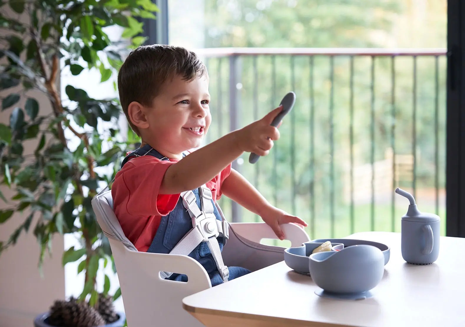 Smiling toddler in a high chair enjoying a meal at the table with natural light and toddler-safe utensils.